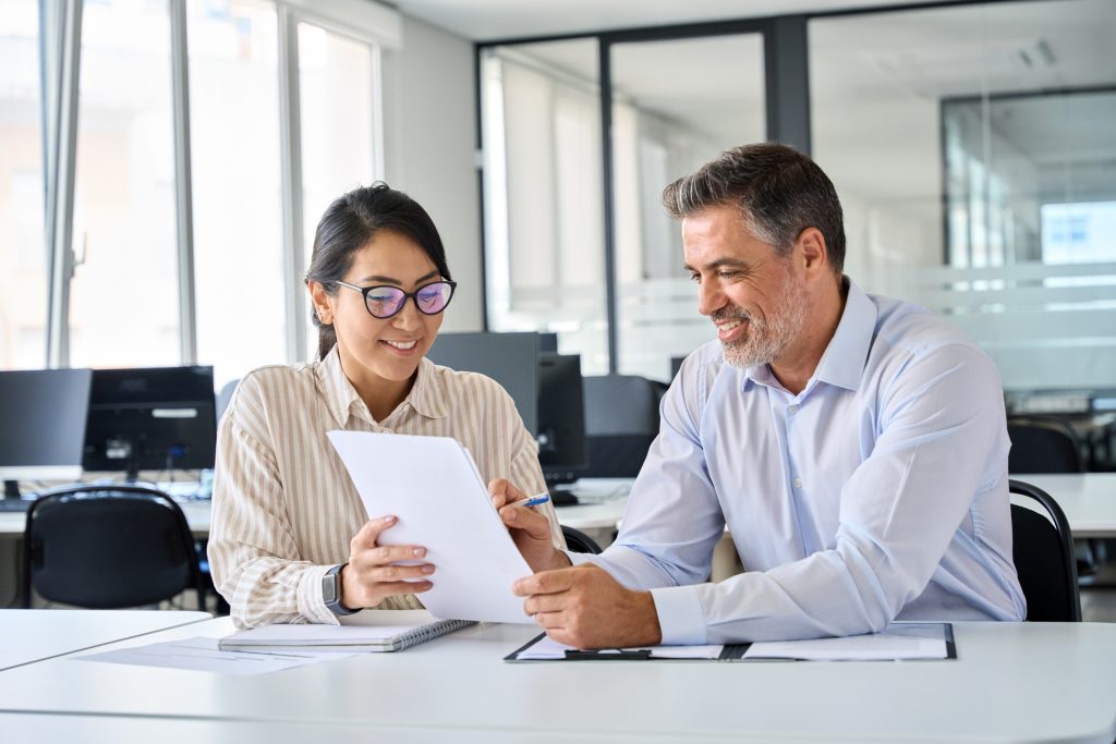 Two professionals reviewing paperwork together at a desk in a modern office, smiling as they discuss documents and take notes.