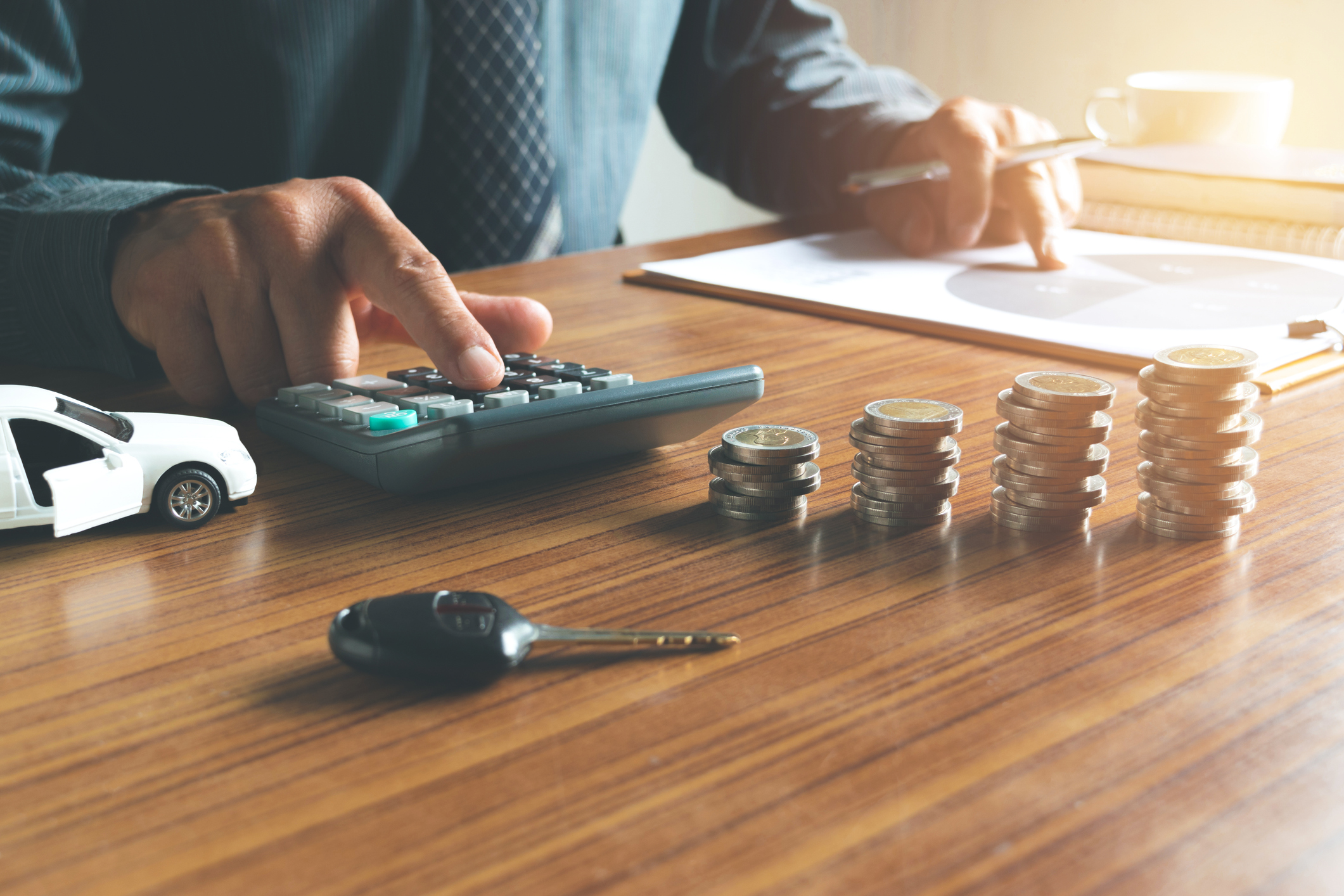 Person calculating car-related expenses using a calculator at a wooden desk, with stacked coins, car keys, a toy car, and financial paperwork in the background.