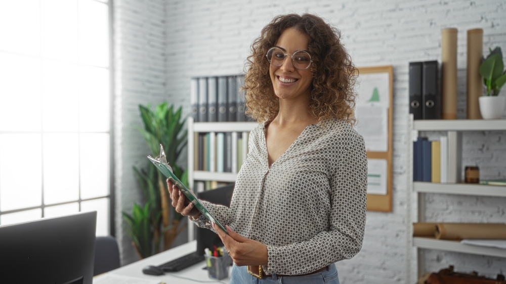 Hispanic woman smiling in modern office holding clipboard surrounded by shelves, plants, and window light creating a professional, relaxed atmosphere showcasing confident, young, Latino businesswoman.
