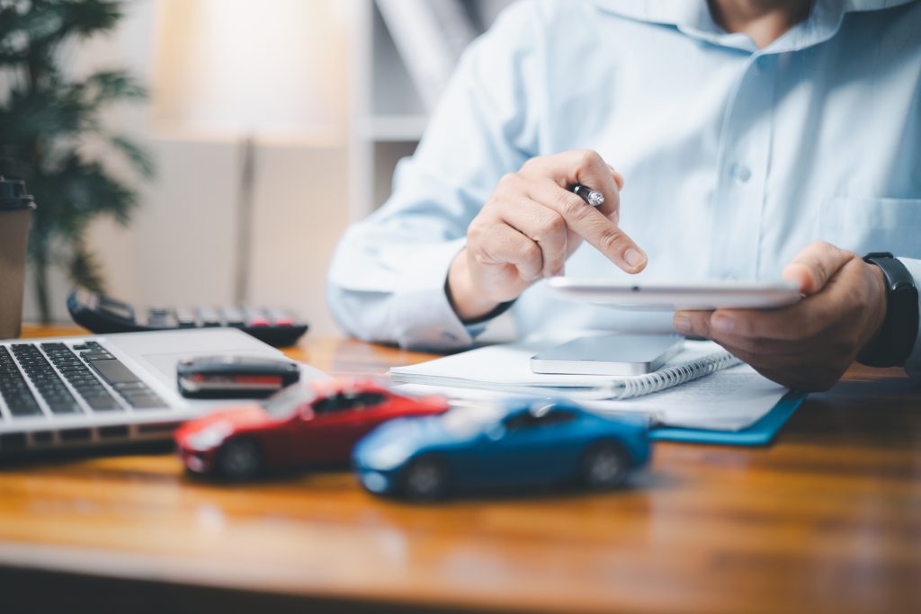 Business professional using a tablet at a desk with model cars, laptop, and paperwork, representing digital tools and operations within a car insurance franchise office.
