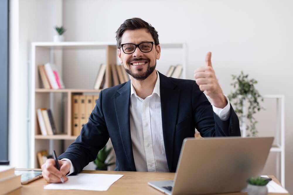 Smiling insurance agent giving a thumbs-up at his desk with a laptop, highlighting how insurance agents make money.
