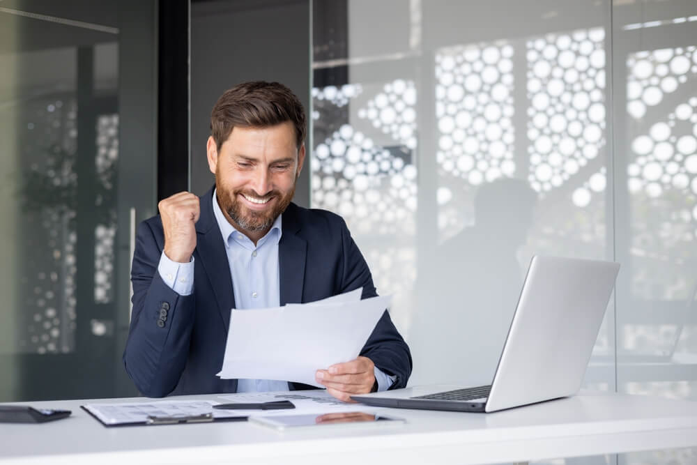 Smiling insurance professional gives a thumbs-up at his desk while reviewing performance reports about how insurance agents make money.