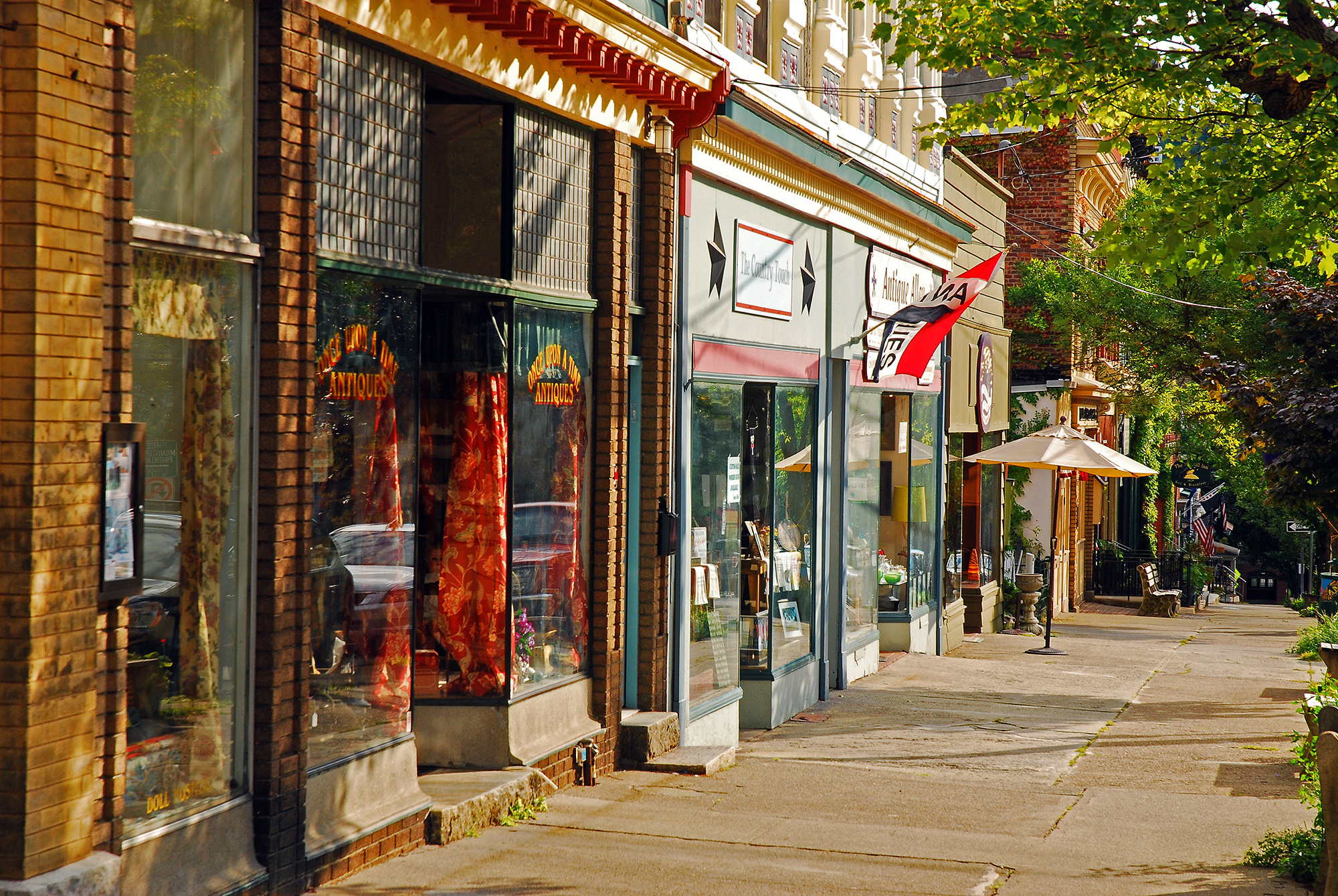 Sunlit small-town street with historic brick storefronts, antique shop windows, flags, and outdoor café umbrellas lining a quiet sidewalk shaded by trees.