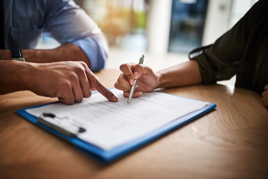 Close-up of two people reviewing and signing a contract at a desk, with one person pointing to the document while the other signs on a clipboard.