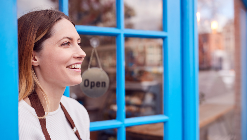 Smiling Female Small Business Owner Standing In Shop Doorway On Local High Street