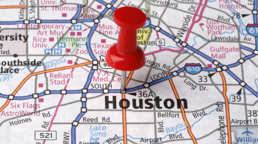 A close-up of a printed map with a red pushpin marking the city of Houston, showing surrounding roads, highways, and landmarks.