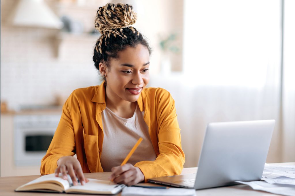 Woman with braided hair in a bun working at a laptop at home, writing notes in a notebook at a kitchen table.