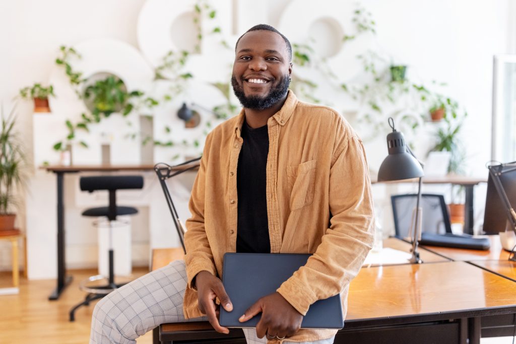 Portrait of a smiling African businessman holding laptop leaning on a coworking desk in office. Male entrepreneur at startup office looking at camera.