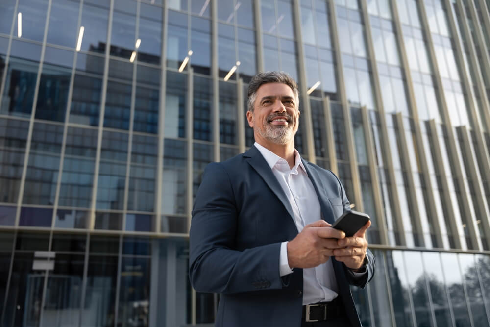 Smiling insurance agent holding a smartphone outside a modern office building, illustrating how insurance agents make money.