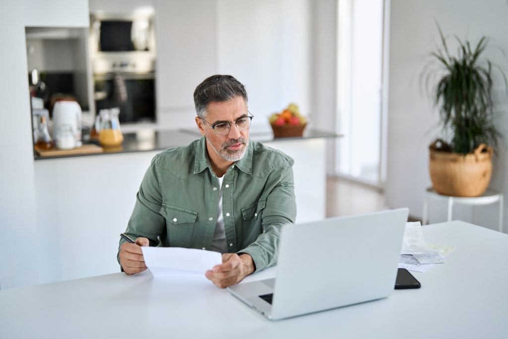 Empresario revisando documentos financieros frente a su computadora portátil en casa, analizando costos y evaluando una oportunidad de franquicia antes de invertir.