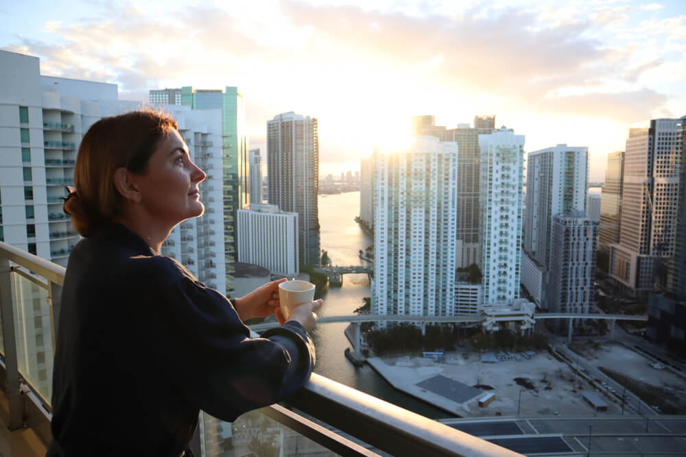 Mujer en balcón con vista urbana al amanecer, representando oportunidades de negocio en Miami.