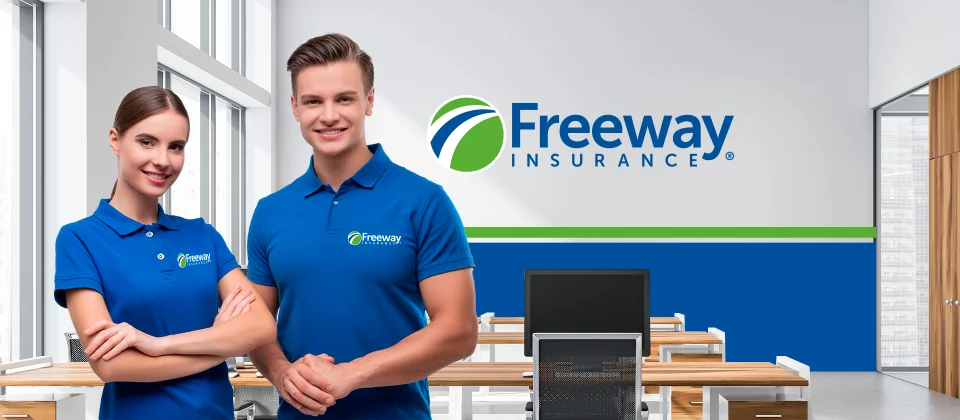 A smiling Anglo man and a woman wearing blue Freeway Insurance brand shirts stand at the back of a Freeway Insurance office smiling at the camera.