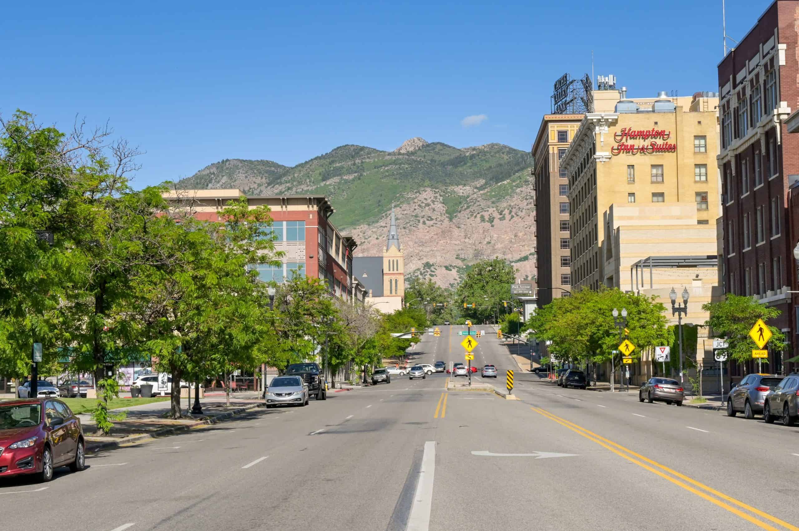 Downtown street in Ogden, Utah, with historic buildings, light traffic, and the Wasatch Mountains rising in the background.
